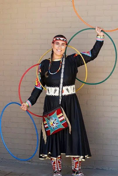 Woman wearing a black dress with beadwork on her dress and moccasins. She is holding four colorful hoops while standing in front of a tan brick wall.