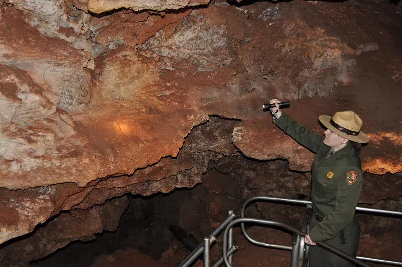 On the right side of the photo is a woman ranger in a green uniform and tan ranger hat using a flashlight to illuminate a red cave wall in the middle of the photo with historic signatures written in black on the wall.