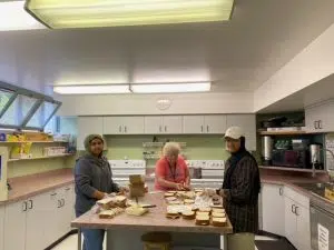 three women making sandwiches in a kitchen stand around a table with lots of bread